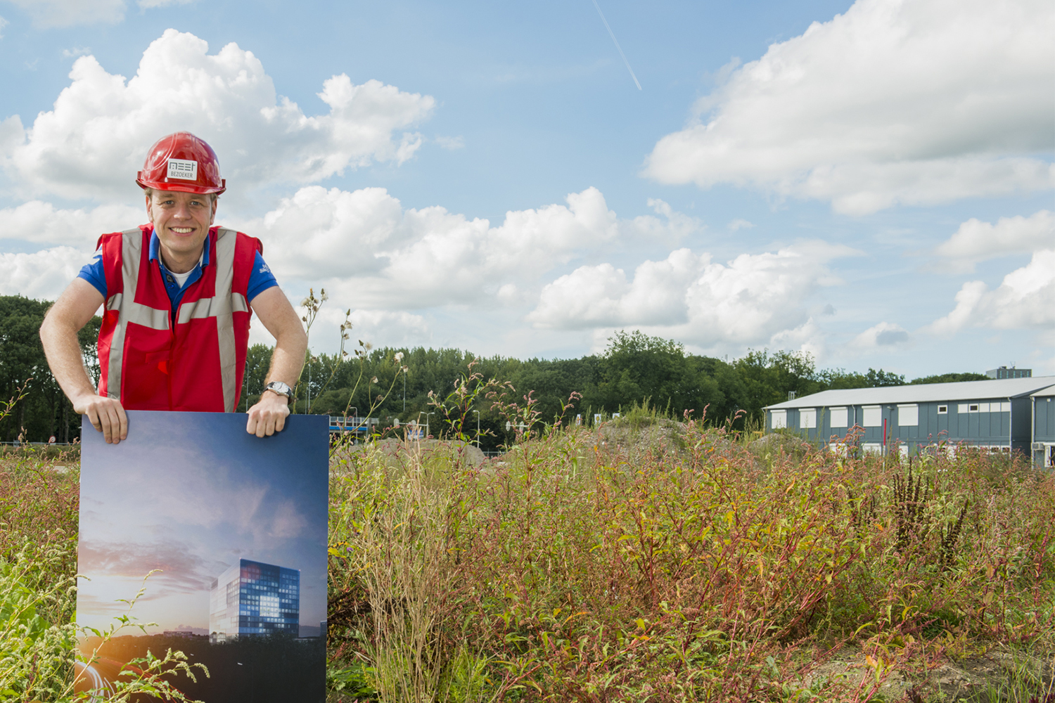 RIVM employee is standing on the new RIVM building site holding a picture with an artist rendering of the new RIVM building.