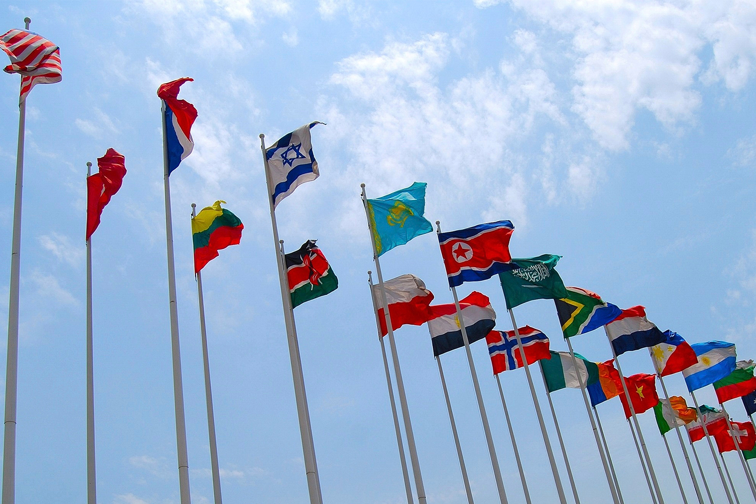 Diverse nationale vlaggen naast elkaar tegen een blauwe lucht met wolken.
